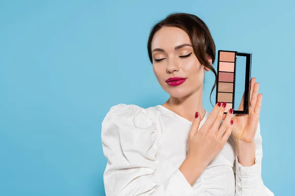 Brunette jeune femme avec les yeux fermés tenant ombre à paupières palette isolé sur bleu — Photo de stock