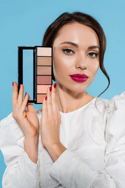 Jolie jeune femme en chemisier blanc tenant palette ombre à paupières isolée sur bleu — Photo de stock