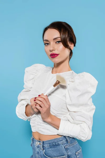 Bonita mujer joven en blusa blanca sosteniendo cepillo cosmético aislado en azul - foto de stock