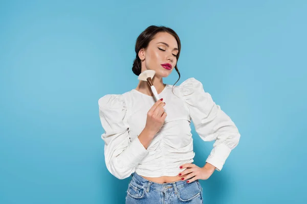 Junge Frau in weißer Bluse berührt Wange mit Kosmetikpinsel und steht mit der Hand auf Hüfte isoliert auf blauem Grund — Stockfoto