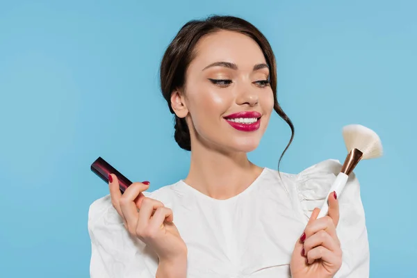 Pretty young woman in white blouse holding cosmetic brush and lipstick isolated on blue — Stock Photo