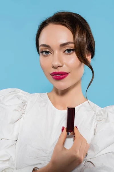 Charming young woman in white blouse holding lipstick isolated on blue — Stock Photo