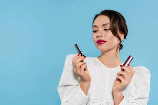 Charming young woman in white blouse looking at lipstick isolated on blue — Stock Photo