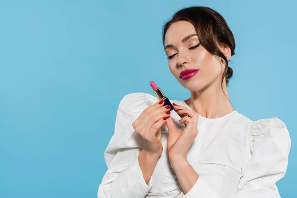 Encantadora joven con los ojos cerrados en blusa blanca sosteniendo lápiz labial aislado en azul - foto de stock