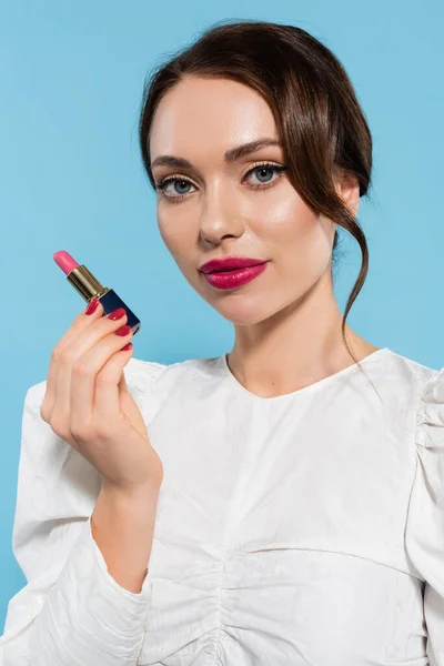 Charming young woman in white blouse holding pink color lipstick isolated on blue — Stock Photo