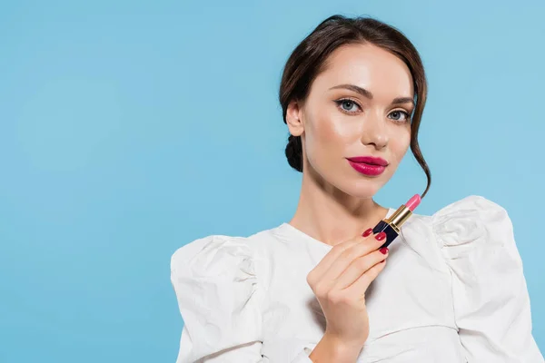 Morena joven en blusa blanca sosteniendo lápiz labial rosa aislado en azul - foto de stock