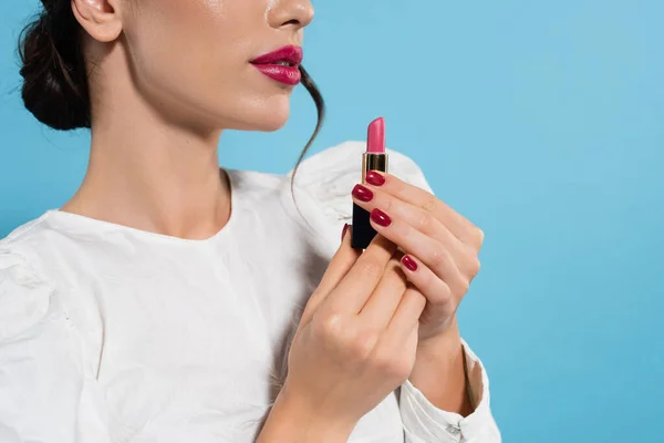 Vista recortada de mujer joven en blusa blanca sosteniendo lápiz labial de color rosa aislado en azul - foto de stock