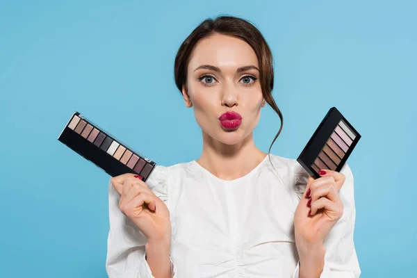 Pretty young woman holding eye shadow palettes and pouting lips isolated on blue — Stock Photo