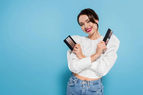 Heureuse jeune femme en chemisier tenant palettes ombre à paupières sur fond bleu — Photo de stock