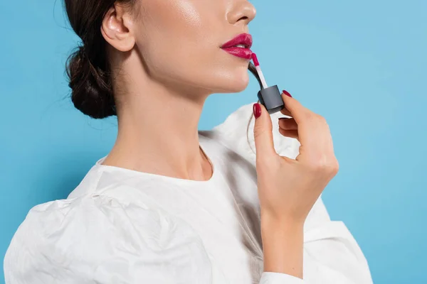 Vista cortada de mulher jovem em blusa aplicando brilho labial rosa no fundo azul — Fotografia de Stock