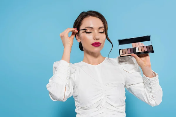 Jolie jeune femme en chemisier blanc appliquant ombre à paupières tout en tenant palette sur bleu — Photo de stock