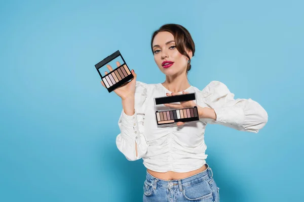 Pretty young woman in white blouse holding shadow palettes in hands on blue background — Stock Photo