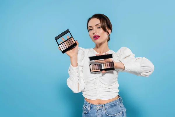 Jolie jeune femme en chemisier blanc tenant des palettes d'ombre dans les mains sur fond bleu — Photo de stock