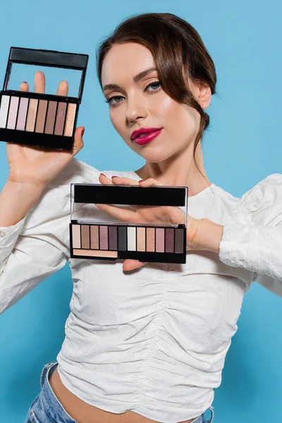 Jolie jeune femme en chemisier blanc tenant des palettes d'ombre dans les mains sur bleu — Photo de stock