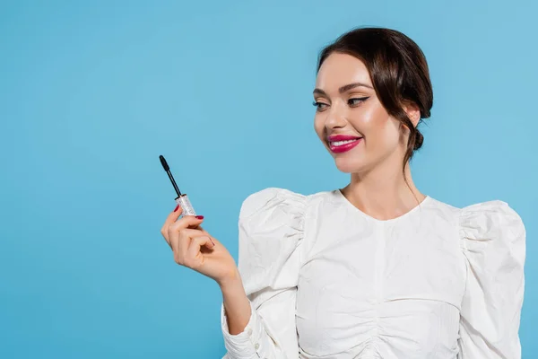 Cheerful young woman in white blouse holding mascara brush isolated on blue — Stock Photo