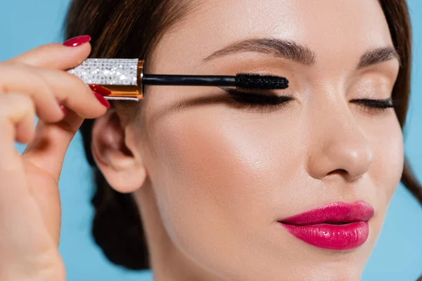 Close up of pretty young woman applying mascara on eyelash isolated on blue — Stock Photo