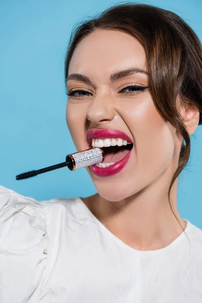 Brunette young woman in white blouse holding mascara brush in teeth isolated on blue — Stock Photo