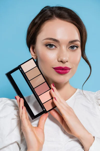 Retrato de bela jovem mulher em blusa branca segurando blush paleta e olhando para a câmera isolada no azul — Fotografia de Stock