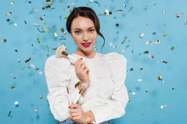 Hübsche junge Frau in weißer Bluse hält Kosmetikpinsel in der Nähe fallenden Konfetti auf blau — Stockfoto