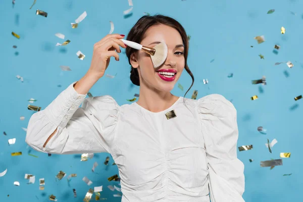 Весела молода жінка в білій блузці, що закриває очі косметичним пензлем біля падаючого конфетті на синьому — стокове фото