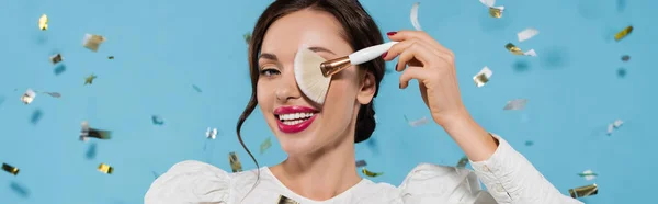 Alegre joven en blusa blanca que cubre el ojo con cepillo cosmético cerca de caer confeti en azul, bandera - foto de stock