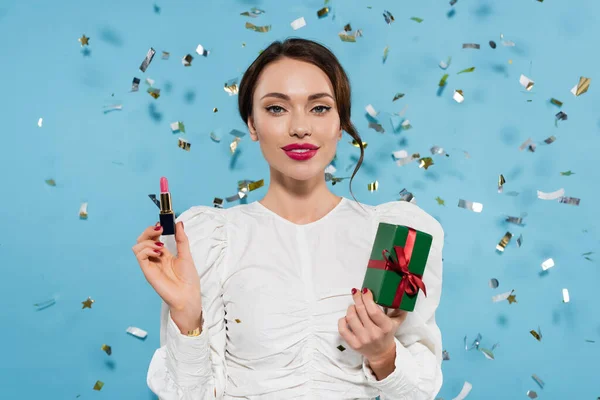 Cheerful young woman in white blouse holding lipstick and gift box near falling confetti on blue — Stock Photo