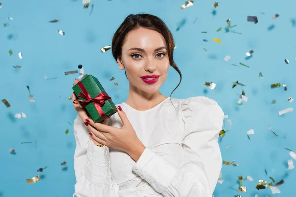 Cheerful young woman in white blouse holding gift box near falling confetti on blue — Stock Photo