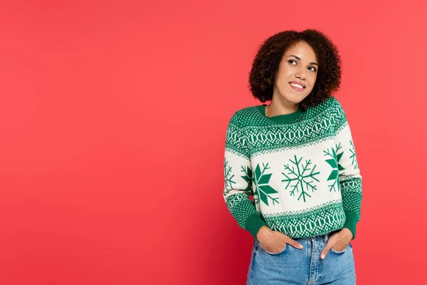 Donna afroamericana sognante e sorridente in maglione invernale in posa con le mani in tasche di jeans isolati su rosso — Foto stock