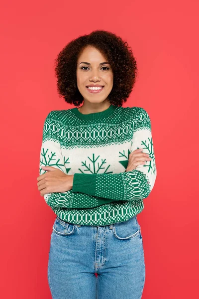 Alegre mujer afroamericana en suéter blanco con patrón verde de pie con brazos cruzados aislados en rojo - foto de stock