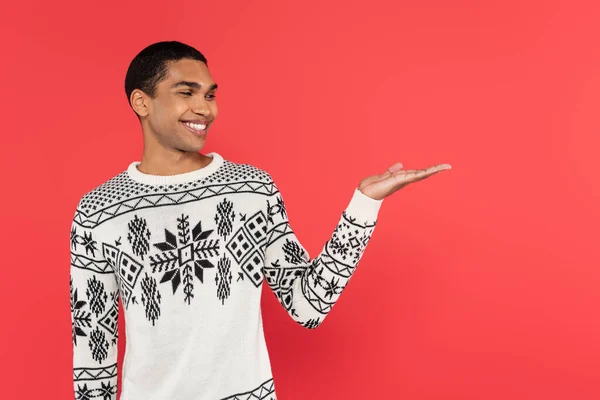 Smiling african american man in knitted winter sweater with ornament pointing with hand isolated on red — Stock Photo