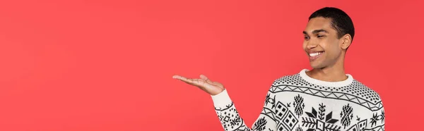 Cheerful african american man in white sweater with winter pattern pointing with hand isolated on red, banner — Stock Photo