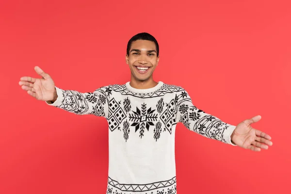 Homem americano africano alegre em camisola quente com ornamento de inverno mostrando gesto de boas-vindas isolado no vermelho — Fotografia de Stock