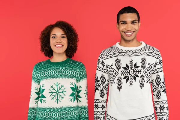 Alegre casal afro-americano em suéteres com ornamento de inverno sorrindo para a câmera isolada no vermelho — Fotografia de Stock