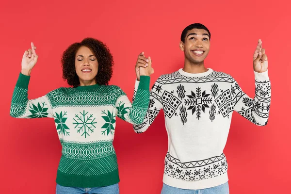 African american couple in sweaters with winter pattern holding hands and crossed fingers isolated on red — Stock Photo