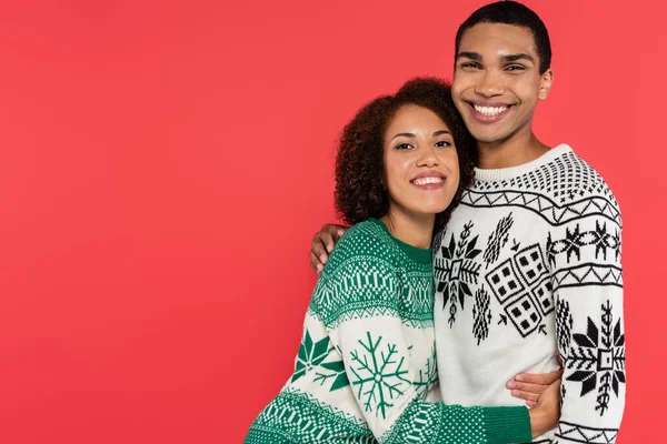 Jovem casal afro-americano em camisolas com padrão de inverno abraçando e sorrindo para a câmera isolada no vermelho — Fotografia de Stock