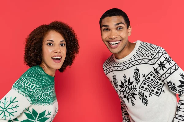 Animado casal afro-americano em camisolas de inverno com ornamento olhando para a câmera isolada no vermelho — Fotografia de Stock