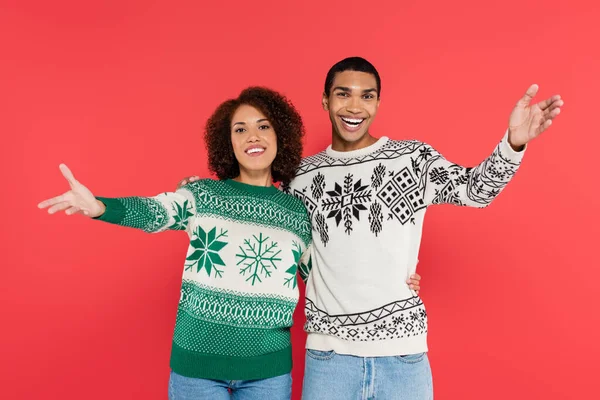 Happy african american couple in warm sweaters with winter ornament showing greeting gesture isolated on red — Stock Photo