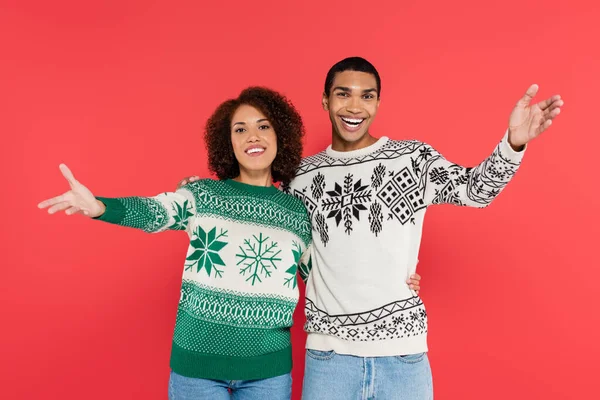 Feliz pareja afroamericana en suéteres cálidos con adorno de invierno mostrando gesto de saludo aislado en rojo - foto de stock