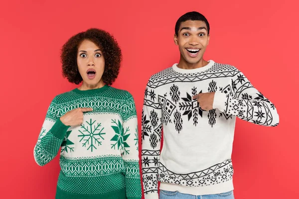 Surprised african american couple looking at camera while pointing with fingers at themselves isolated on red — Stock Photo