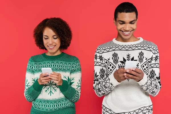 Smiling african american couple in sweaters with winter pattern chatting on smartphones isolated on red — Stock Photo