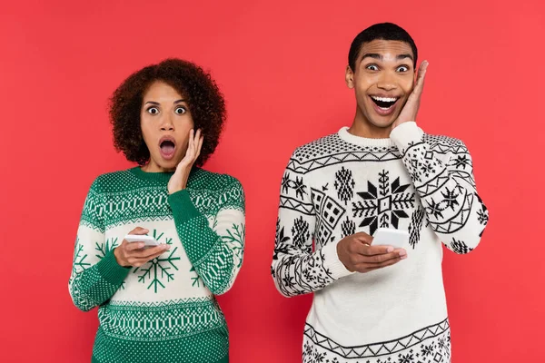 Pareja afroamericana sorprendida con teléfonos móviles mirando a la cámara aislada en rojo - foto de stock