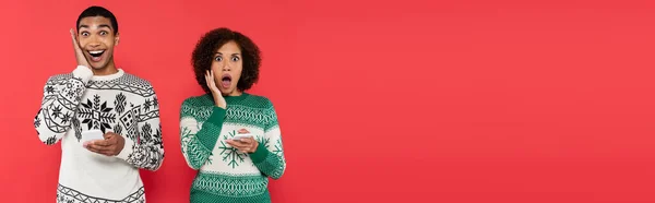 Donna afroamericana scioccata in maglioni invernali che tengono i telefoni cellulari e guardando la fotocamera isolata su rosso, banner — Foto stock