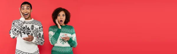 Impactó a la mujer afroamericana en suéteres de invierno sosteniendo teléfonos celulares y mirando a la cámara aislada en rojo, pancarta - foto de stock