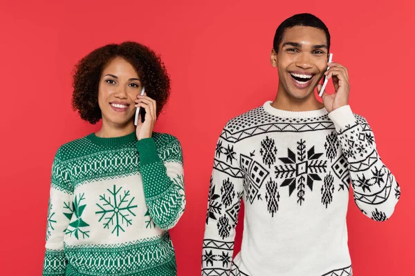 Sonriente pareja afroamericana en suéteres cálidos con patrón de invierno hablando en teléfonos inteligentes aislados en rojo - foto de stock