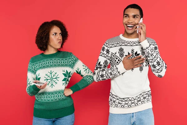 Offended african american woman standing with hand on hip near laughing boyfriend talking on smartphone isolated on red — Stock Photo