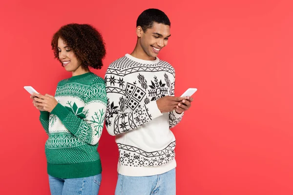 Heureux couple afro-américain debout dos à dos et la messagerie sur les smartphones isolés sur rouge — Photo de stock