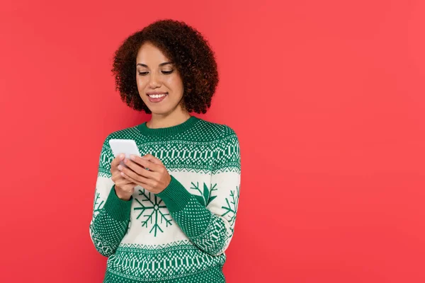 Positive brunette african american woman in white sweater with green ornament using smartphone isolated on red — Stock Photo