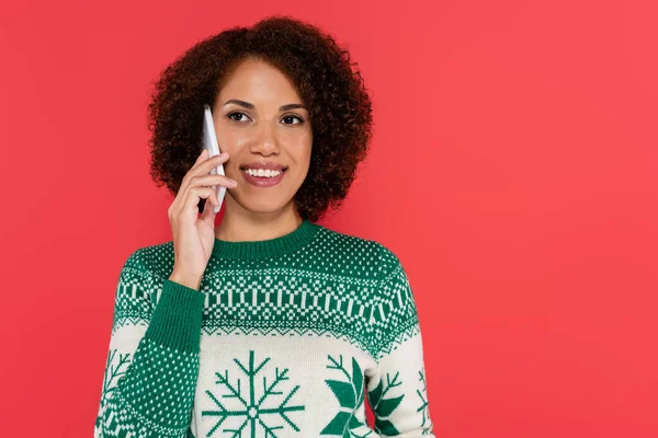 Femme afro-américaine bouclée en pull d'hiver avec motif vert parlant sur téléphone portable isolé sur rouge — Photo de stock