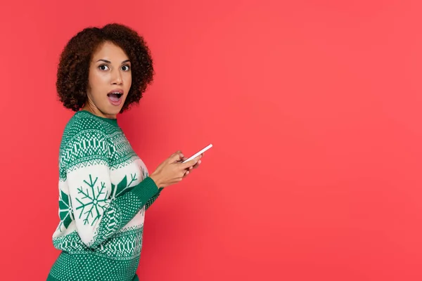 Amazed african american woman in warm sweater holding smartphone and looking at camera isolated on red — Stock Photo