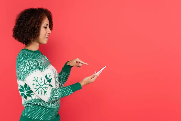 Side view of brunette african american woman in warm sweater pointing with finger at smartphone isolated on red — Stock Photo